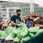 A person wearing a mask and gloves sorts green bags in front of piles of cardboard boxes at an outdoor distribution site.