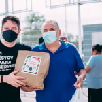 Two men wearing face masks stand outdoors; one holds a paper bag with a holiday-themed label. Other people are visible in the background.