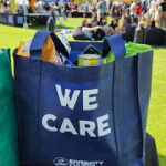 A blue tote bag with "WE CARE" printed on it, filled with various items, sits on grass at an outdoor event with people in the background.