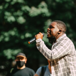 A man holding a microphone speaks outdoors, whilst another person wearing a mask stands in the background.