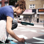 A person crouches on a city street painting blue and white patterns on the pavement, with shops visible in the background.