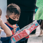 A young boy wearing a black face mask looks at a red packaged item whilst standing outdoors beside another child.