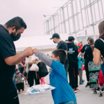 A man and a child fist bump outside near a crowd of people; the child holds a parcel while others stand and walk in the background.