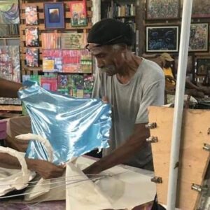 Three people work together in an art studio, stretching and attaching a sheet of blue material to a frame, surrounded by artwork and supplies.