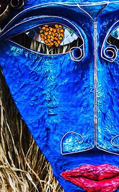 A close-up of a vibrant blue decorative mask with wire detailing, textured golden hair, and bright red lips. One eye features a cluster of small orange shapes.