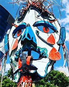 Large, colourful abstract sculpture of a human face with exaggerated features, metal accents, and a blue sky with trees in the background.