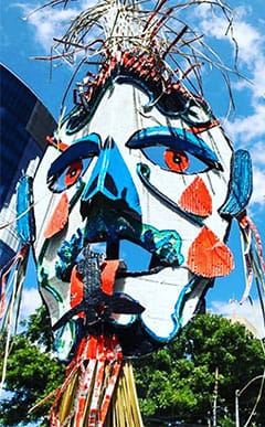 Large, colourful abstract sculpture of a human face with exaggerated features, metal accents, and a blue sky with trees in the background.
