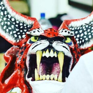 A person paints a large, red, horned mask with sharp fangs, wide open mouth, and pointed ears, resembling a fierce, mythical creature.