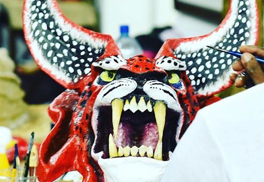 A person paints a large, red, horned mask with sharp fangs, wide open mouth, and pointed ears, resembling a fierce, mythical creature.