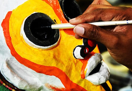 A person paints the eye of a vibrant, yellow-orange papier-mâché mask with a paintbrush.