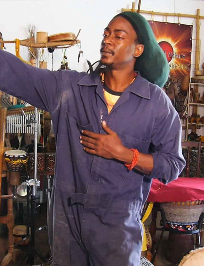 A man in a blue boiler suit and green hat stands indoors with one hand on his chest, surrounded by musical instruments and cultural artefacts.