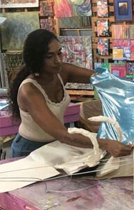 A woman in a vest works with shiny blue material in an art studio filled with colourful paintings and art supplies.