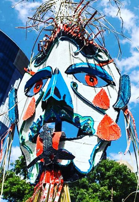 Large colourful sculpture of an abstract human face made from painted materials, displayed outdoors against a blue sky and trees.